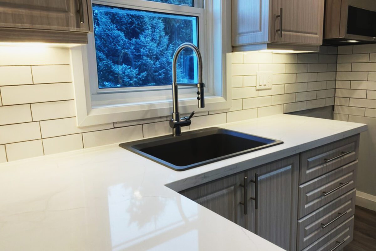 Looking an 'L' shaped counter to a black sink and kitchen facet in from of a window.
